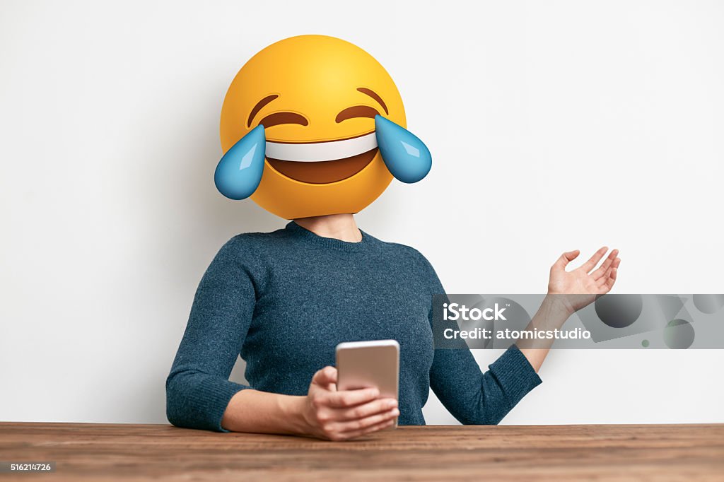 Emoji Head Woman sitting at desk. Emoji Head Woman sitting at desk. Woman wearing tears of joy emoji masks while looking at her phone. This emoji is laughing so much that it is crying tears of joy Emoticon Stock Photo