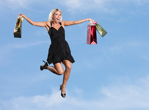 A beautiful young caucasian woman dressed in a black skirt with heels and holding shopping bags jumps with excitement. Mid air with sky and clouds in the background.