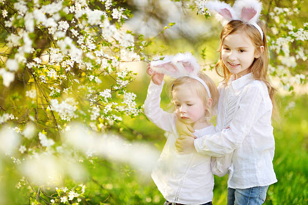 Two little sisters wearing bunny ears on Easter Two adorable little sisters wearing bunny ears in a spring garden on Easter day holding child flower april stock pictures, royalty-free photos & images