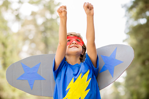 Latin descent, cute little boy plays superhero outdoors on his neighborhood street in spring or summer season.  He has made a costume for himself with mask and airplane wings.  He pretends to \