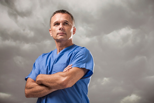Candid portrait of a handsome, mature male doctor looking off into the distance with a serious, somewhat worried expression. Set against a moody, ominous cloudy sky background.