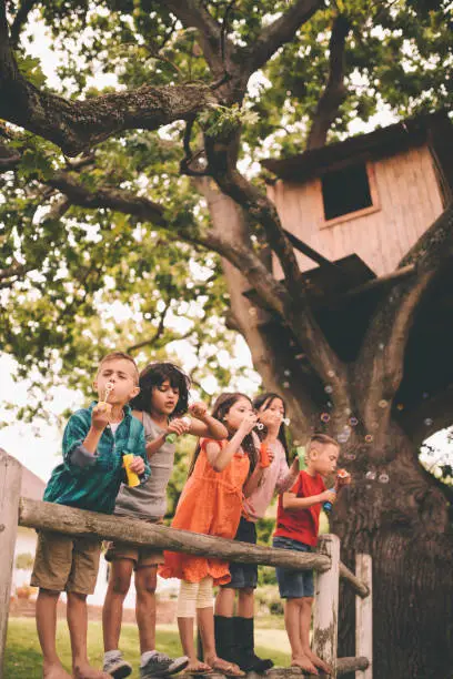 Long haired hispanic boy laughing and having fun with his friends standing on a wooden fence in a summer park blowing bubbles, with a vintage develop