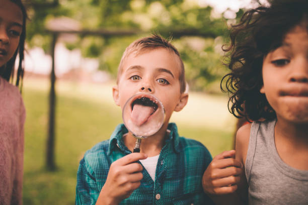 Little boy in a park with friends holding a large round magnifying glass up to his mouth while sticking out his tongue, making it look ridiculously huge