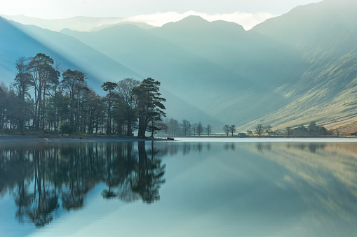 Beautiful sunbeams shining through a thin layer of mist with calm reflections of trees in the lake at Buttermere in the English Lake District. Taken on a spring morning, the green colours are starting to appear on the mountain sides.