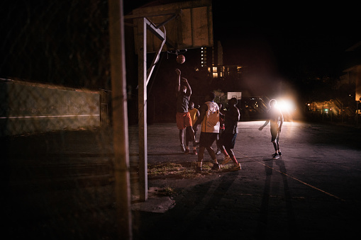 African American basketball players playing in inner-city court with man jumping to dunk ball into basket during nighttime with single court light in background