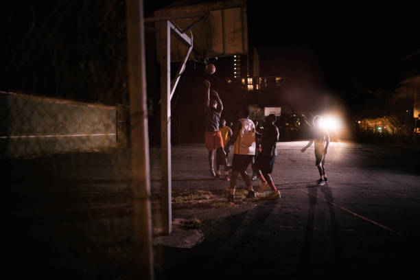 Basketball Players Playing in Court During Nighttime African American basketball players playing in inner-city court with man jumping to dunk ball into basket during nighttime with single court light in background silhouette of basketball player slam dunk stock pictures, royalty-free photos & images
