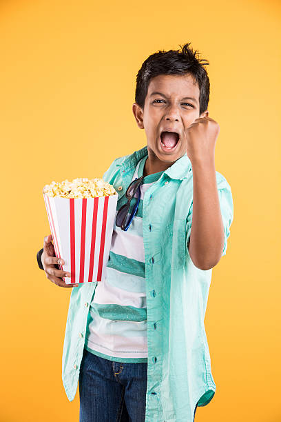 Joyful indian kid with big box of popcorn, eating popcorn Joyful indian kid holding a big box of popcorn and looking at the camera isolated on yellow background, Portrait of adorable indian young boy eating popcorn, asian kid and popcorn 10 11 years photos stock pictures, royalty-free photos & images