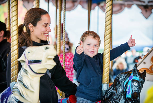 Boy with Two Thumbs Up with Mother on Carousel A happy mother and son are riding on a carousel together, smiling and having fun at an amusement park.  The boy holds two thumbs up. carnival children stock pictures, royalty-free photos & images