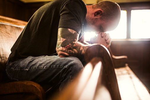 A man bows his head in prayer while sitting on a wooden pew in a traditional church building. Horizontal image with copy space.