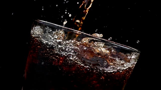 Cola soda pouring into glass of ice with splashes at slow motion on a black background