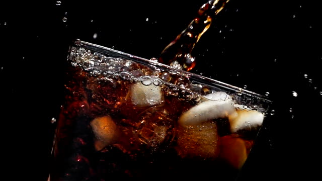 Cola soda pouring into glass of ice with splashes at slow motion on a black background