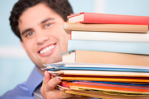 Happy Latin descent business man or college student holding large stack of files, books, paperwork.  Close up.  Office, home or school. 