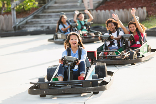 Multi-ethnic group of teens (16 to 18 years) riding go carts at amusement park.  Focus girl in front.