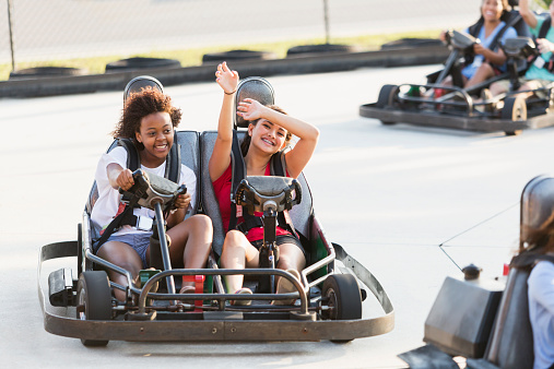 Multi-ethnic teenage girls riding go carts at amusement park.  Focus on two girls in back (16 years).