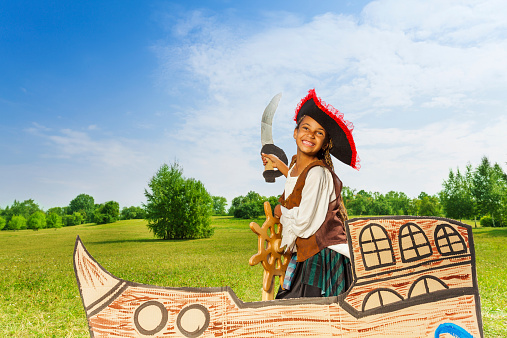 Happy African girl in pirate costume with hat and sword holds helm on carton ship