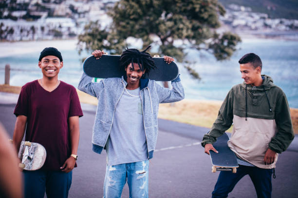 Multi-Ethnic group of skater friends hanging out, holding skateboards and smiling with one holding skateboard behind head with both hands at seaside