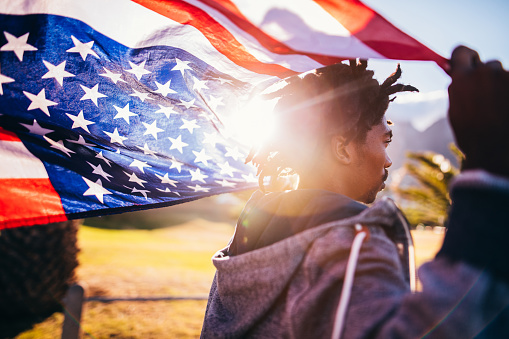 African American skater holds an American flag behind his head while walking outside during sunset