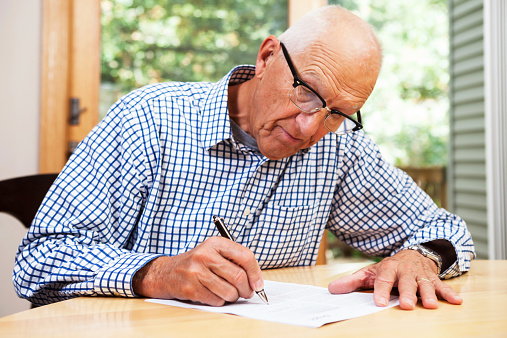 A senior man, in his 70s, is sitting at a dining room table filling out paperwork and signing a document.