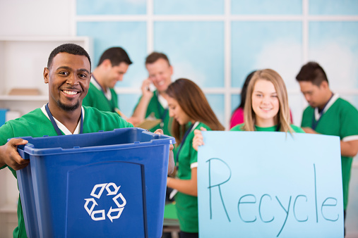 Multi-ethnic group of young adults collaborate to organize a recycling campaign.  African descent man foreground with recycling bin. Female volunteer holds \