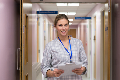 A portrait shot of an young female doctor standing in a hospital corridor looking at the camera and smiling. She is holding a clipboard and looks like she is on her way to do the rounds.