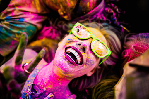 Close Up face shot of girl covered in colorful powder smiling and making peace sign while laying down on grass in a park during Holi Festival