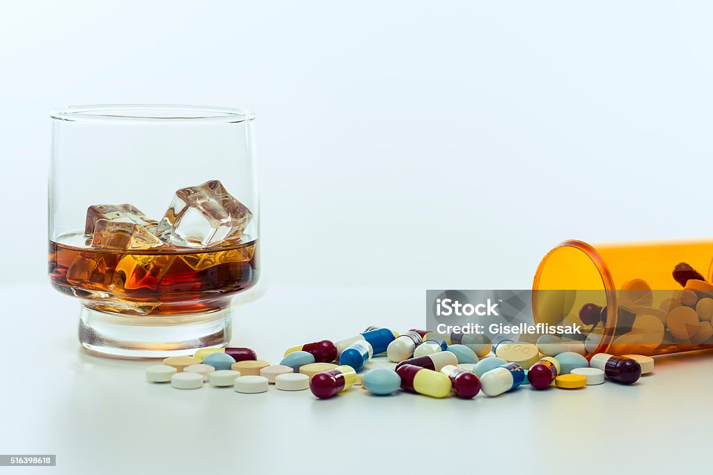 Alcohol and drugs. Whiskey glass, with ice cubes, beside a large amount of drugs, capsules and pills spread around and falling out of the recipient on a white background. Medicine Stock Photo