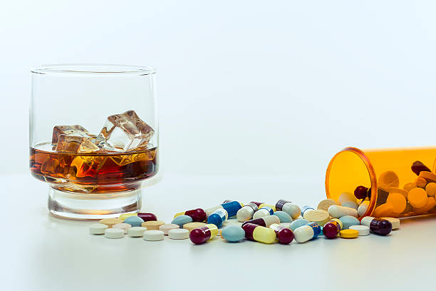 Alcohol and drugs. Whiskey glass, with ice cubes, beside a large amount of drugs, capsules and pills spread around and falling out of the recipient on a white background. substance abuse stock pictures, royalty-free photos & images