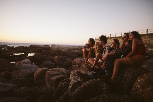 Side view of a group of young hipster friends enjoying a sunset at the sea sitting on rocks