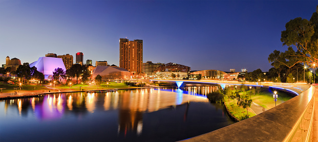 brightly lit Adelaide city CBD with foot bridge across Torrens river. Illumination reflecting in calm waters at sunrise.