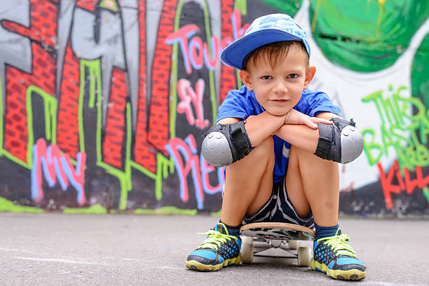 Attractive young boy in a trendy blue outfit sitting on his skateboard in front of a brightly colored graffiti wall smiling at the camera