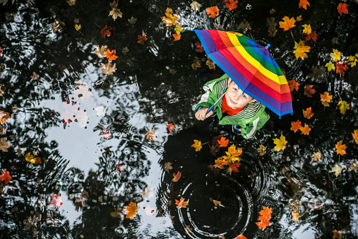 Little boy standing in the water puddle on the rainy day with colorful umbrella and looking up.