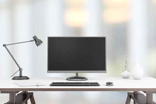 View at a working desk,  with a large PC monitor,lamp, a plant, coffee cup, with a blurry out of focus business background.