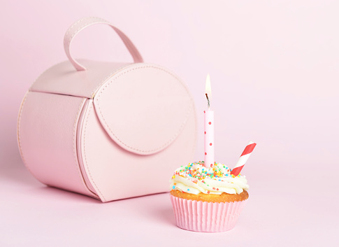 Cupcake, with buttercream flavor and sprinkles on top of sugar cream in a pink paper patty pan on a pink background with a pink birthday candle lit up for a girls party and pink hand bag in background