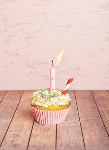 Cupcake, with buttercream flavor and sprinkles on top of sugar cream in a pink paper patty pan with a pink background wall on brown wood slates and a pink birthday candle ontop lit up