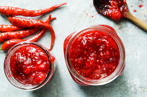 Two jam jars filled with cranberry and red chile jam are ready to be processed for storing.  The image shot directly from above using natural light shows unused red chiles, a wooden spoon and jars of jam. 