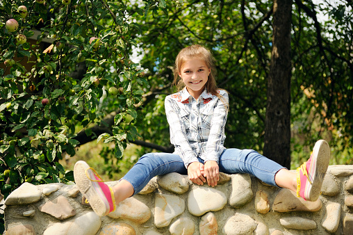 [b]Summer[/b], Preadolescent girl, legs spread, sitting on a wall of stone and smiling cheerfully behind her green trees.