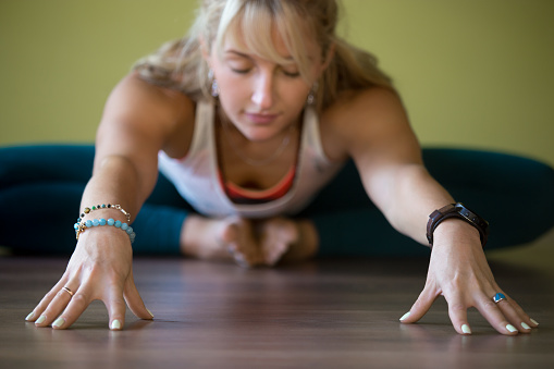 Sporty beautiful blond young woman in sportswear working out indoors, doing Butterfly Pose with closed eyes, sitting in Purna Titli or Baddha Konasana Posture, full length, close-up, focus on hands