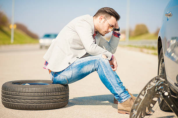 Car problems A young man with a silver car that broke down on the road.He is sitting near car with punctured tire. flat tire stock pictures, royalty-free photos & images