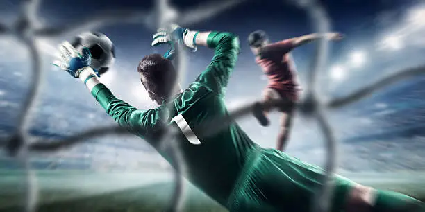 Behind the gate net view - a male soccer goalie jumping in motion for a ball while defending his gates on wide angle panoramic image of a outdoor soccer stadium or arena full of spectators under a sunny sky. The image has depth of field with the focus on the foreground part of the pitch. Players are wearing unbranded soccer uniform.