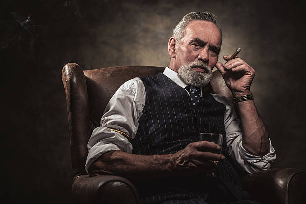 In chair sitting senior business man with cigar and whisky. In chair sitting senior business man with cigar and whisky. Gray hair and beard wearing blue striped gilet and tie. Against brown wall. man cigar stock pictures, royalty-free photos & images