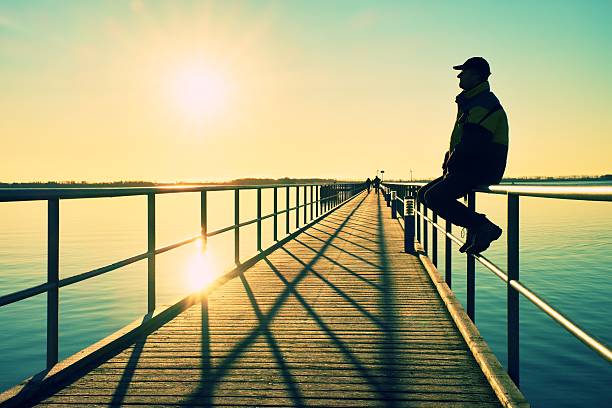 Man in warm jacket and baseball cap sit on pier handrail construction and enjoy morning at sea. Sunny clear blue sky, smooth water level