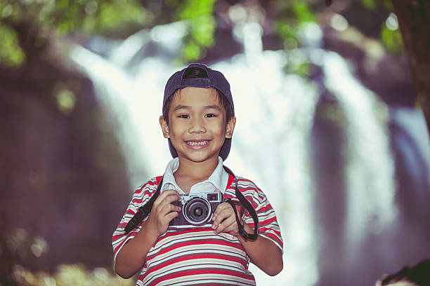 Asian child smiling and holding professional digital camera on blurred waterfall background. Handsome boy in nature. Outdoors portrait. Vintage picture style.