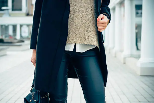 Details of women's clothing. Close-up of a woman in a sweater, coat, black pants. In her hand female bag.