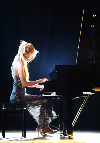 Side view od adult caucasian woman playing piano in a concert. She's placed on a theatre stage. Wearing elegant long dress. Partially backlit, vertical shot. Black stage curtain in background.