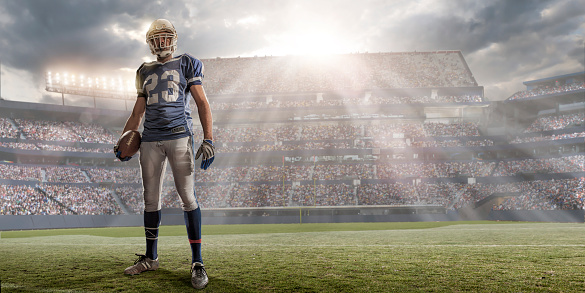 Professional American football player – running back - holding football and standing alone on American football pitch. The player is in a generic outdoor floodlit American football stadium full of spectators under an stormy evening sky at sunset with bright sunlight. Player is wearing generic kit and stadium advertising is fake. 