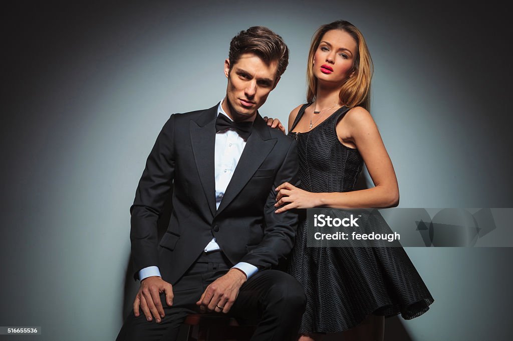 elegant couple in black posing in studio elegant couple in black posing in studio background looking at the camera. man is seated while woman stands by his side with hands on his shoulder and arm Men Stock Photo