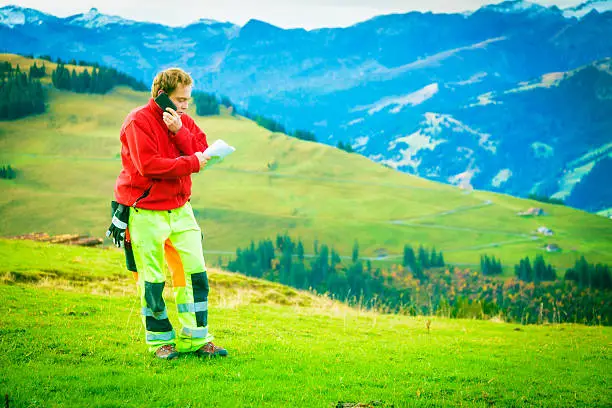 paramedic standing on alpine meadow, talking on the phone and looking through documents.