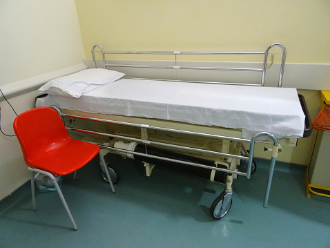 Photo showing a hospital examination table / bed / couch in a doctor's office, ready for the next patient to arrive.  The bed has been covered with a removable white plastic sheet, for hygiene purposes.  A red plastic chair is pictured in the foreground.