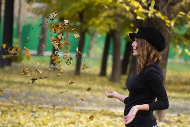 Portrait of an attractive beautiful pretty cute young caucasian cheerful smiling successful woman (girl, female, person, model) dressed in a black peak cap playing with old dry autumn foliage in a park.