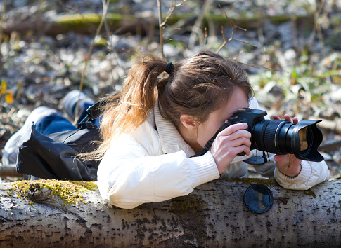 Portrait of photographer (young woman). The girl is hidding to take secretively images of wild life in a spring day sunny forest.